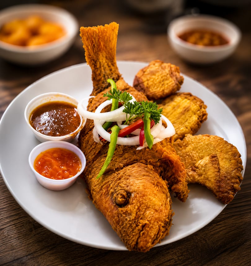 Crispy whole fried fish with onion, peppers, parsley, and two dipping sauces on a white plate.